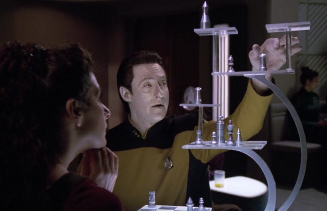 Data and Deanna Troi from Star Trek: The Next Generation are playing 3D chess. Data is reaching for a piece on the board, while Deanna looks on.