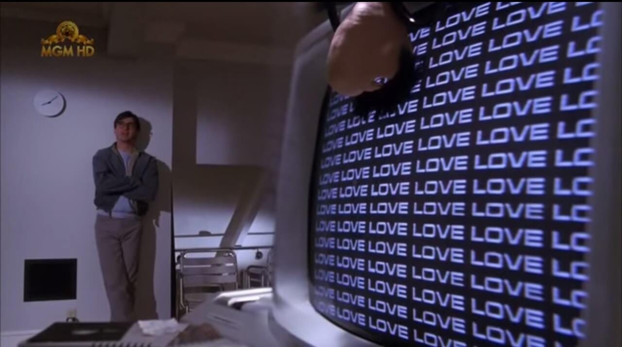 A still from the movie Electric Dreams. An 80s desktop computer with the word LOVE repeatedly on its screen. A man leans against the wall in the background, arms crossed.