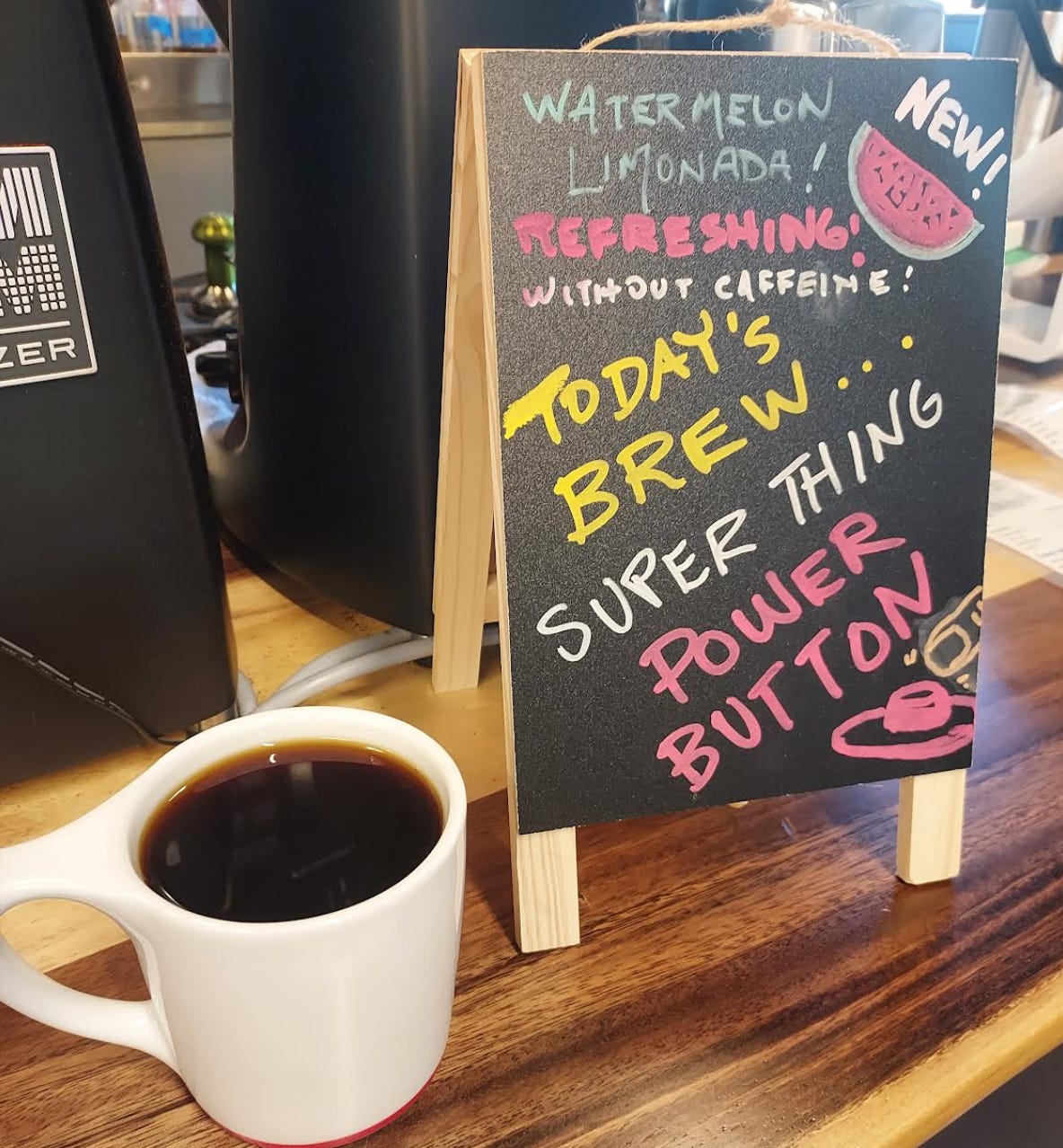 From an above angle: A white coffee cup is full of coffee next to a tabletop chalkboard sign declaring the brew is Super Thing Power Button next to a chalk drawing of a watermelon slice.