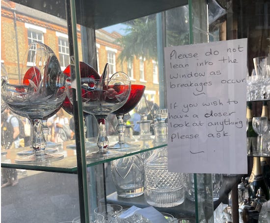 A handwritten sign on a shelf of glass chalices that reads "Please do not lean into the window as breakages occur. If you wish to have a closer look at anything please ask."