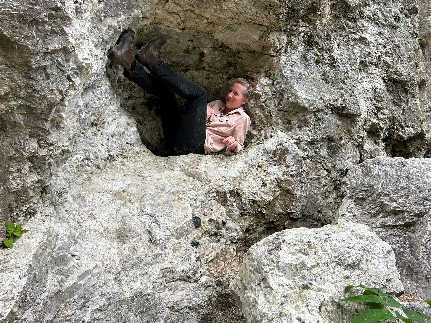 Photo of Michele in cavern in the quarry walls, smiling with messy bun, pink long-sleeve shirt, dirty black jeans, and boots.