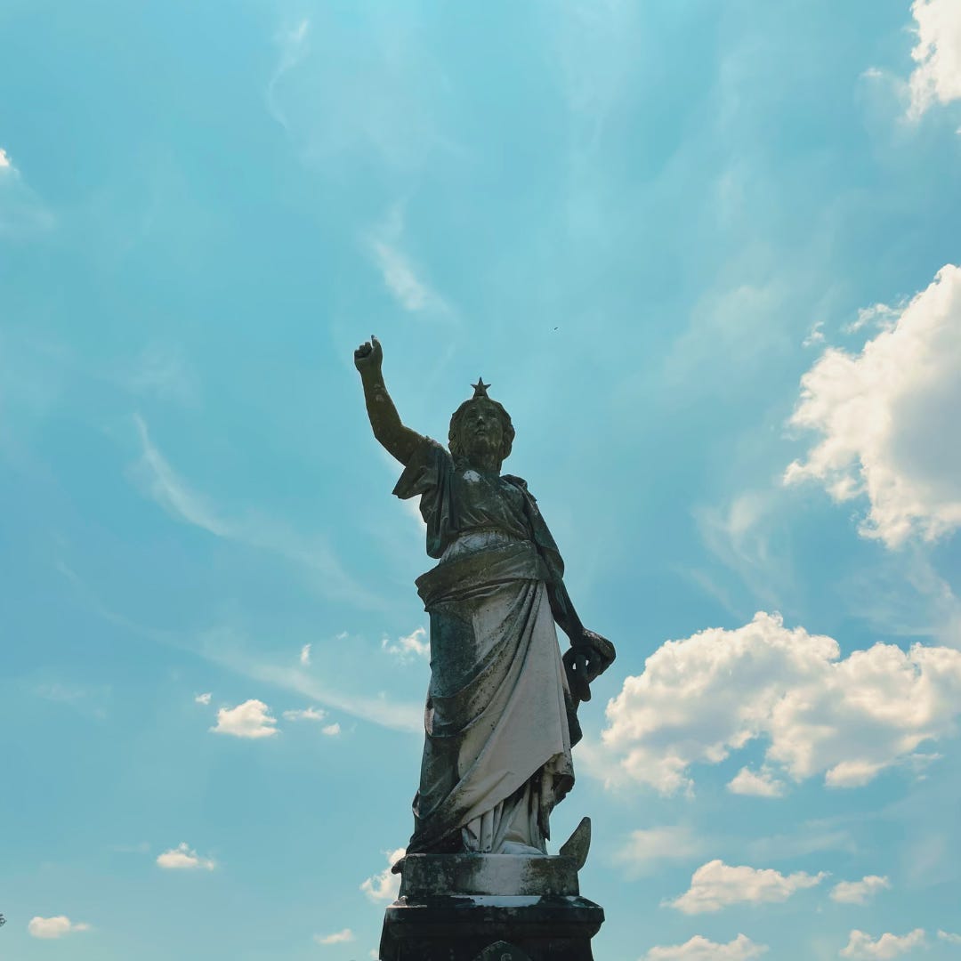 A statue of a woman with one arm raised, on top of a gravestone, and with a bright blue sky in the background.