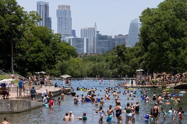 Large numbers of people wade in a pool with trees and skyscrapers in the background.