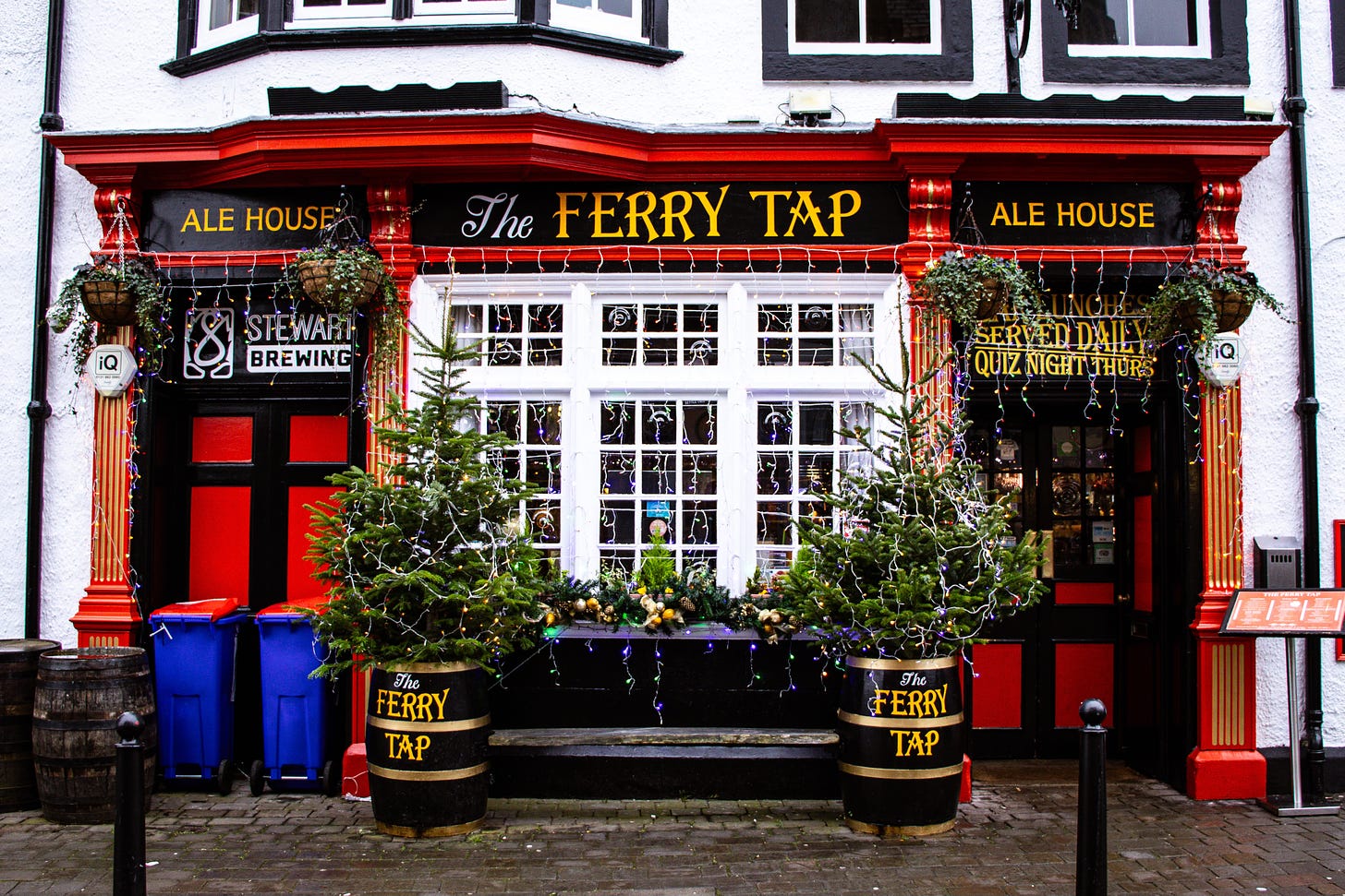 Exterior of the pub, with red painted frontage, yellow sign on black background. Christmas trees and lights in the barrels on the street.