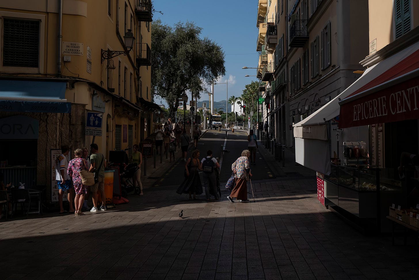 A European street scene with pedestrians walking through a dimly lit alleyway, framed by old buildings and shopfronts, with a glimpse of greenery in the distance.