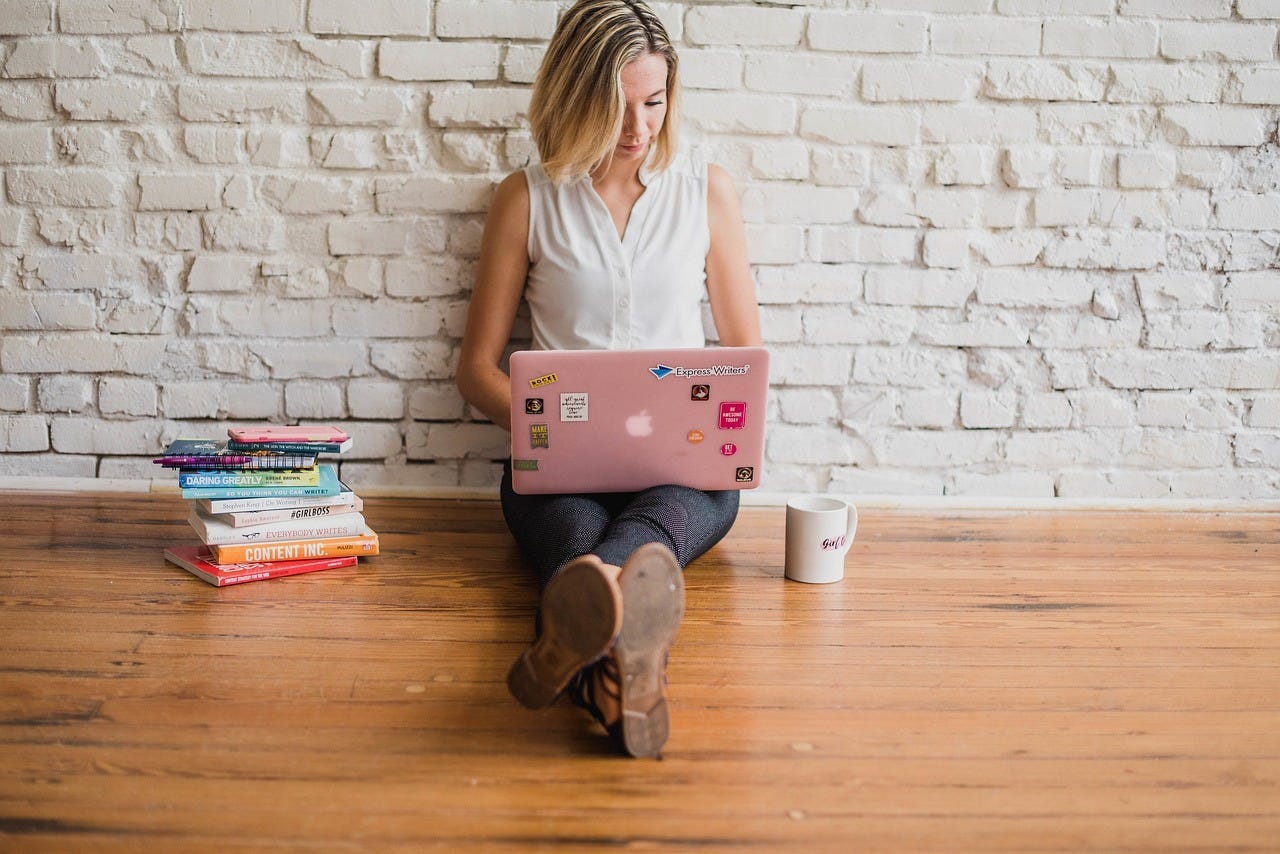 Woman on laptop next to stack of books used to deliver more value to her readers and earn more income. Woman on laptop next to stack of books used to deliver more value to her readers and earn more income.