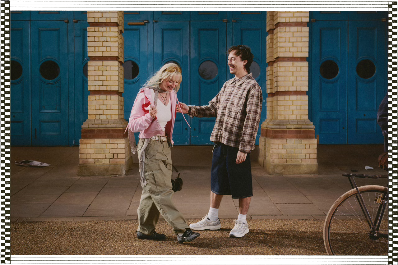 Caitlin and Tian share a laugh outside a brick building with bold blue doors. She wears cargo pants and a pink hoodie with a heart, while he wears a plaid shirt and long shorts. A bicycle wheel appears at the edge of the frame.