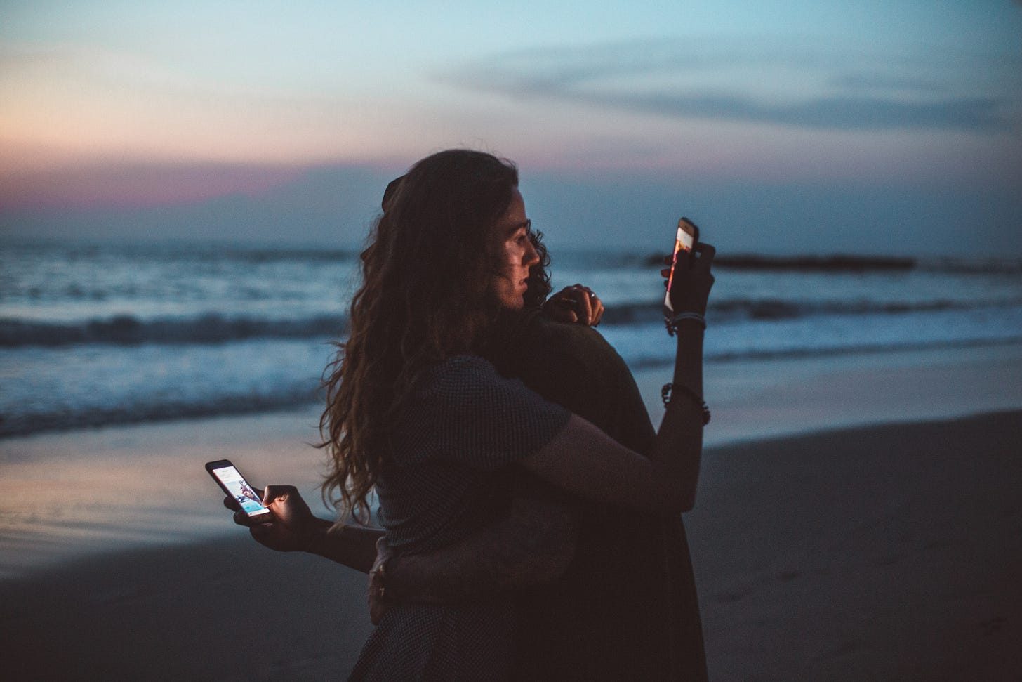 Couple hugging on the beach by the ocean at sundown ignoring the sunset and each other to stare at their phones behind each other's backs.