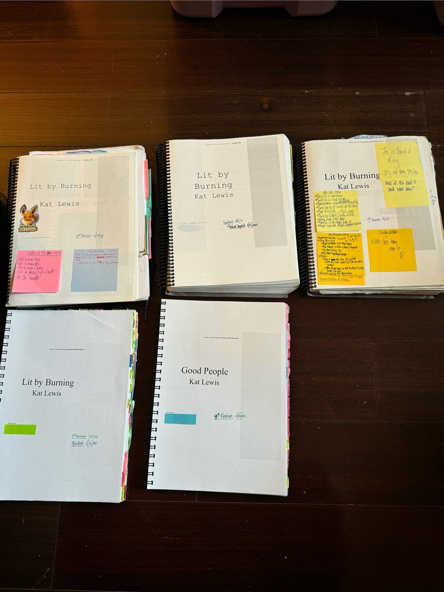 A photo of five printed and bound manuscripts of Kat’s novel, GOOD PEOPLE. Each manuscript is thick, battered, and adorned with a plethora of post-it notes. All but one title page contains the novel’s previous title, LIT BY BURNING. Some of the manuscripts date as far back as June 2018. The most recent manuscript is dated October 2024.