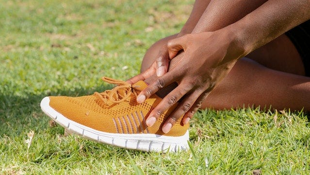 Sitting on the grass, a brown-skinned person holds their ankle—which s n a yellow shoe with a white sole—as if in pain.
