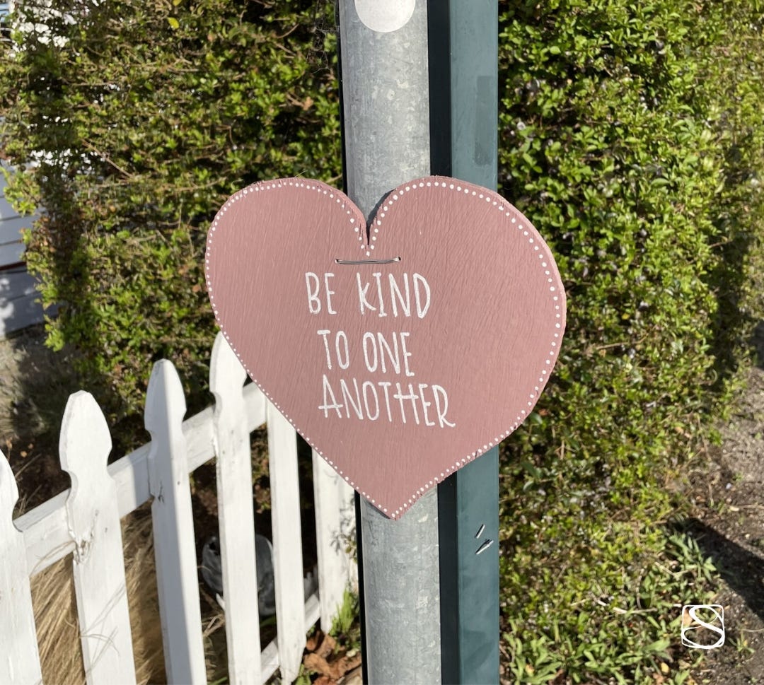 'Be kind to one another' painted on a pink wooden heart hanging from a street pole