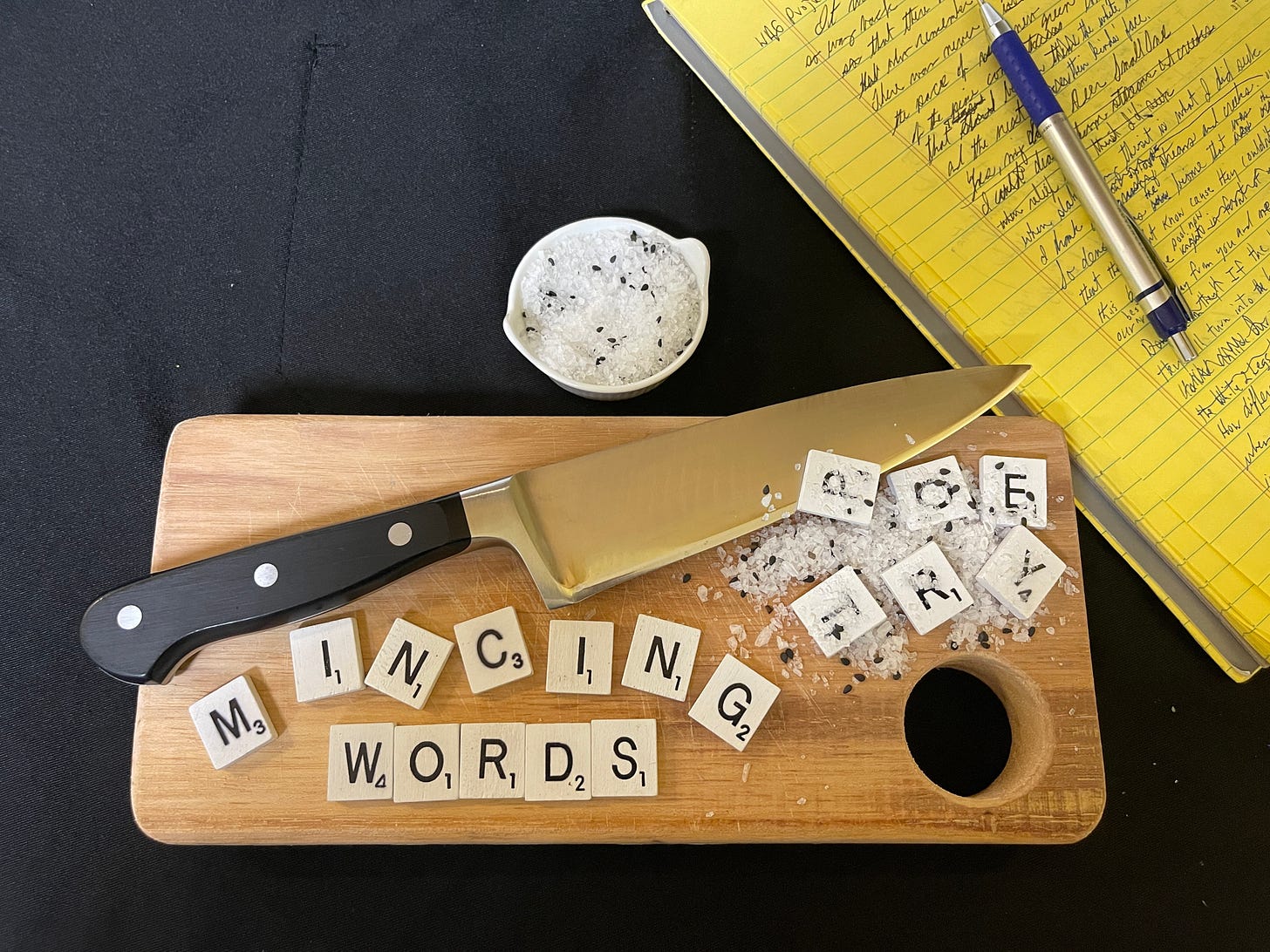 The phrase “mincing words”spelled out in scrabble letters is on the left side of a wood cutting board. A chef’s knife lays diagonally across the cutting board just above the “mincing words” phrase.  The letter “P” lays on part of the knife, while the other scrabble letters that spell out “poetry” are mixed in with a pile of finely chopped (minced) white and black pieces.  In the upper right of the picture is a yellow lined notepad full of prose written out in cursive form and a silver and blue pen laying on the notepad.