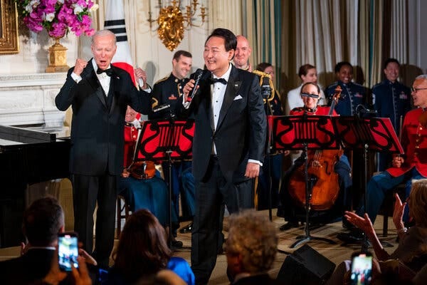 President Biden raises his fists in a cheer as Mr. Yoon holds a microphone and sings before a crowd. Both leaders are wearing tuxedos.