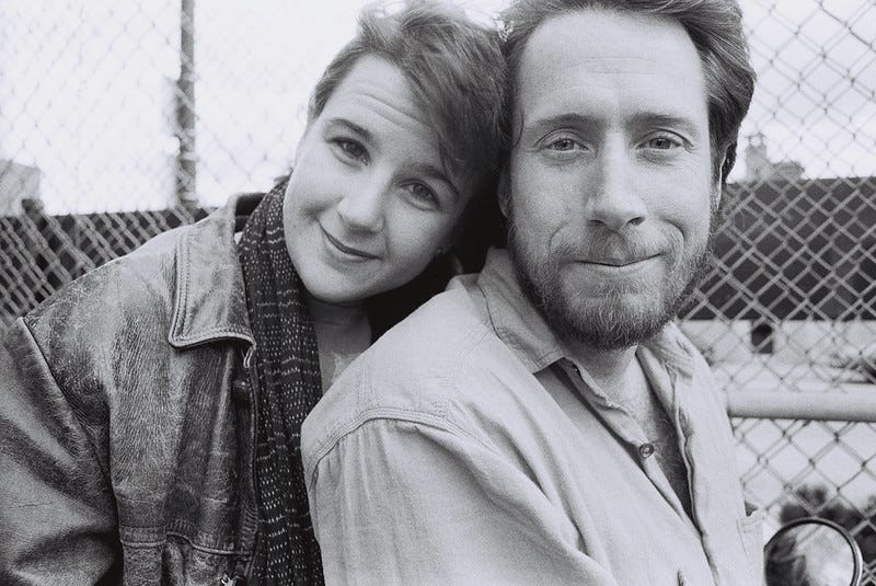 A black-and-white photo of a young woman, wearing a leather jacket and scarf, smiling and leaning her head toward a smiling, bearded man. There is a playground-type fence behind them.