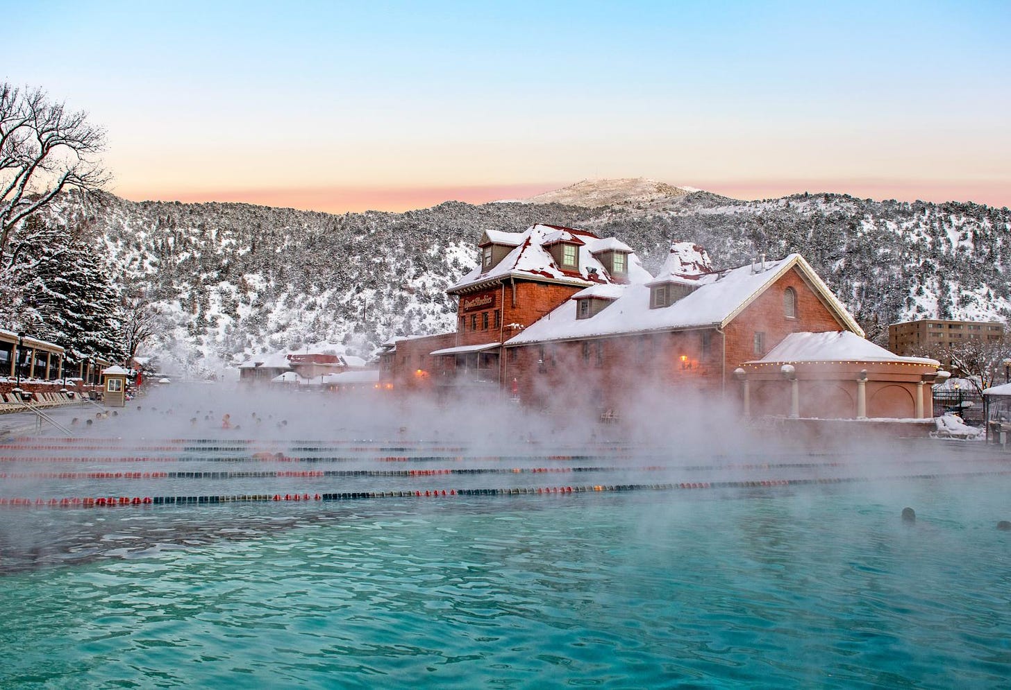 Steam pours off of an enormous outdoor swimming pool, filled with swimmers. In the background, snow covers the buildings around it and the slopes of Glenwood Canyon.