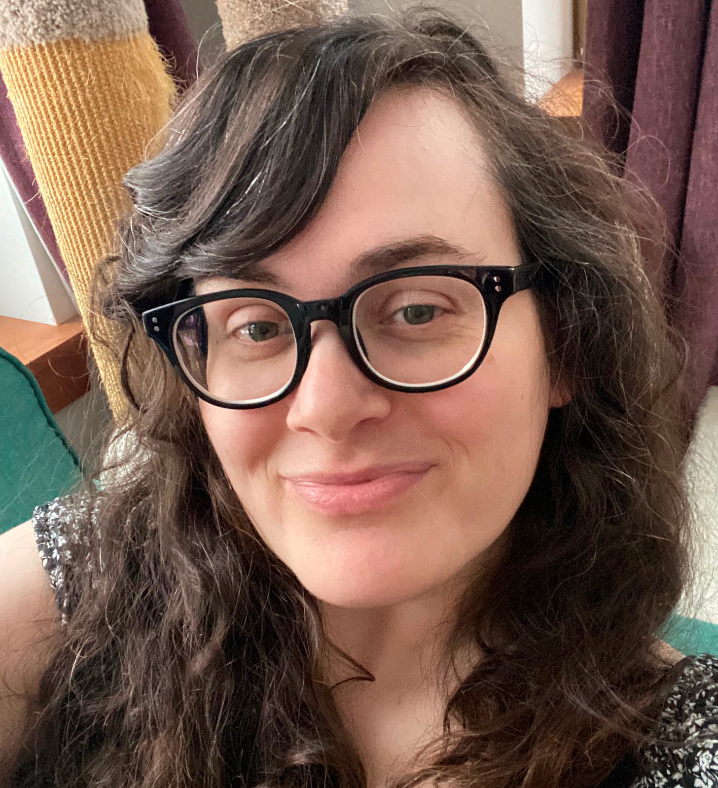 Image description: A picture of Tiffany smiling at the camera.  She is wearing black glasses and a black and white dress.  In the background you can see a green sofa and a well loved cat tree. 