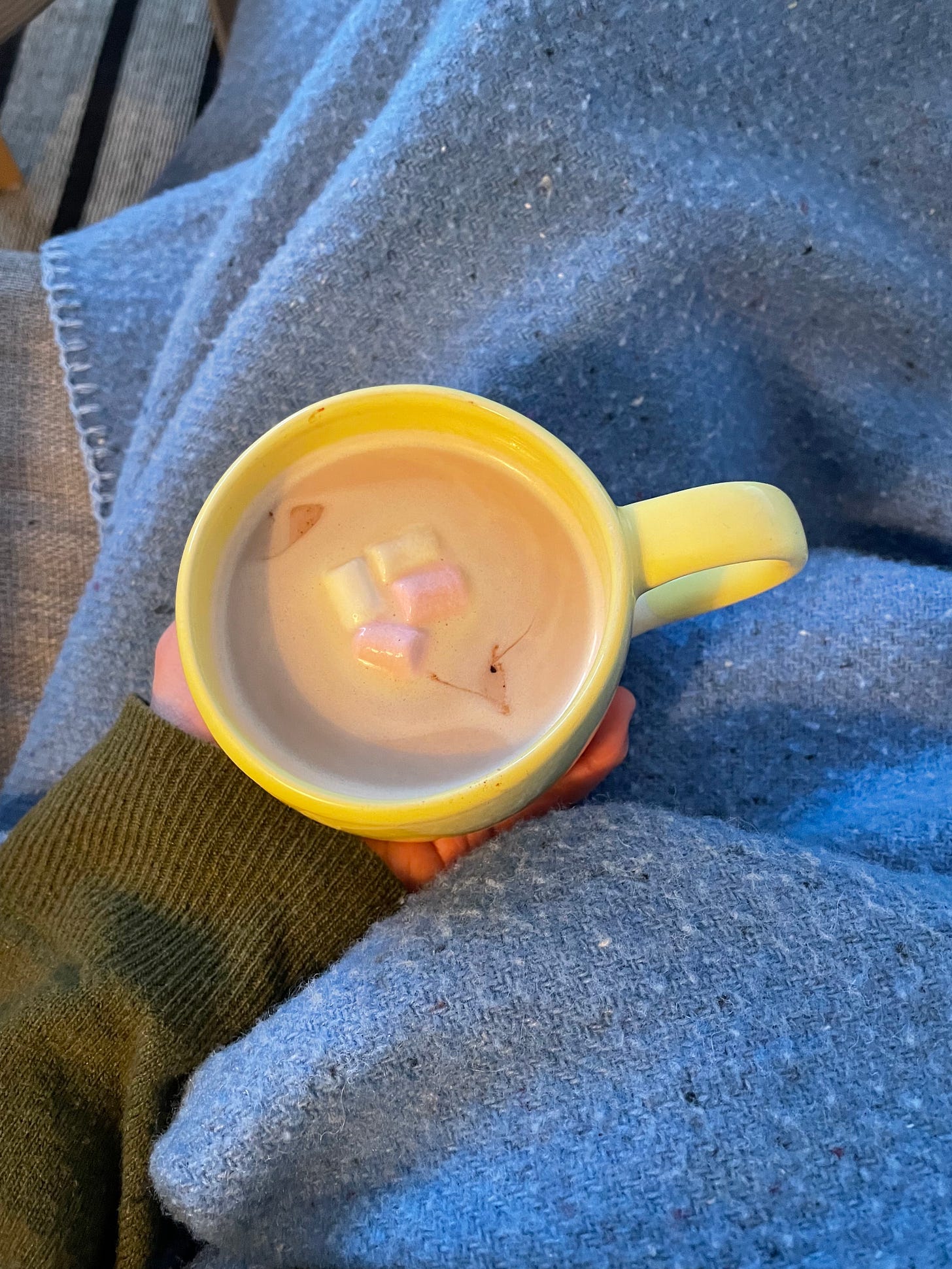 Cup of hot chocolate with melting pink and white mini marshmallows. In the background, a woollen blanket and a rug. A person drinking hot chocolate on the couch, watching TV.