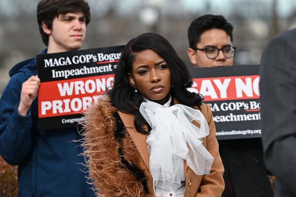 Representative Jasmine Crockett stands in front of two people holding up protest signs referencing the “MAGA GOP.” She is wearing a brown jacket with furry trim and a white shirt with a bow.
