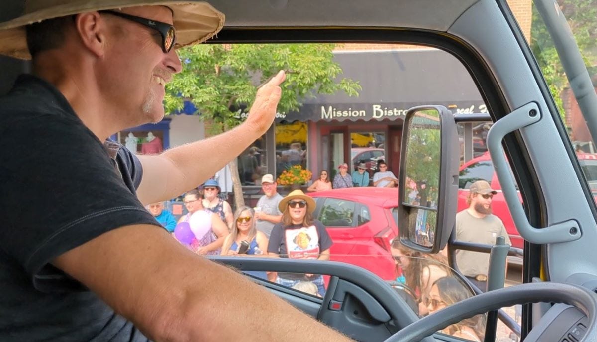 a man in a hat and sunglasses smiling and waving to a crowd outside the window of the truck he is driving