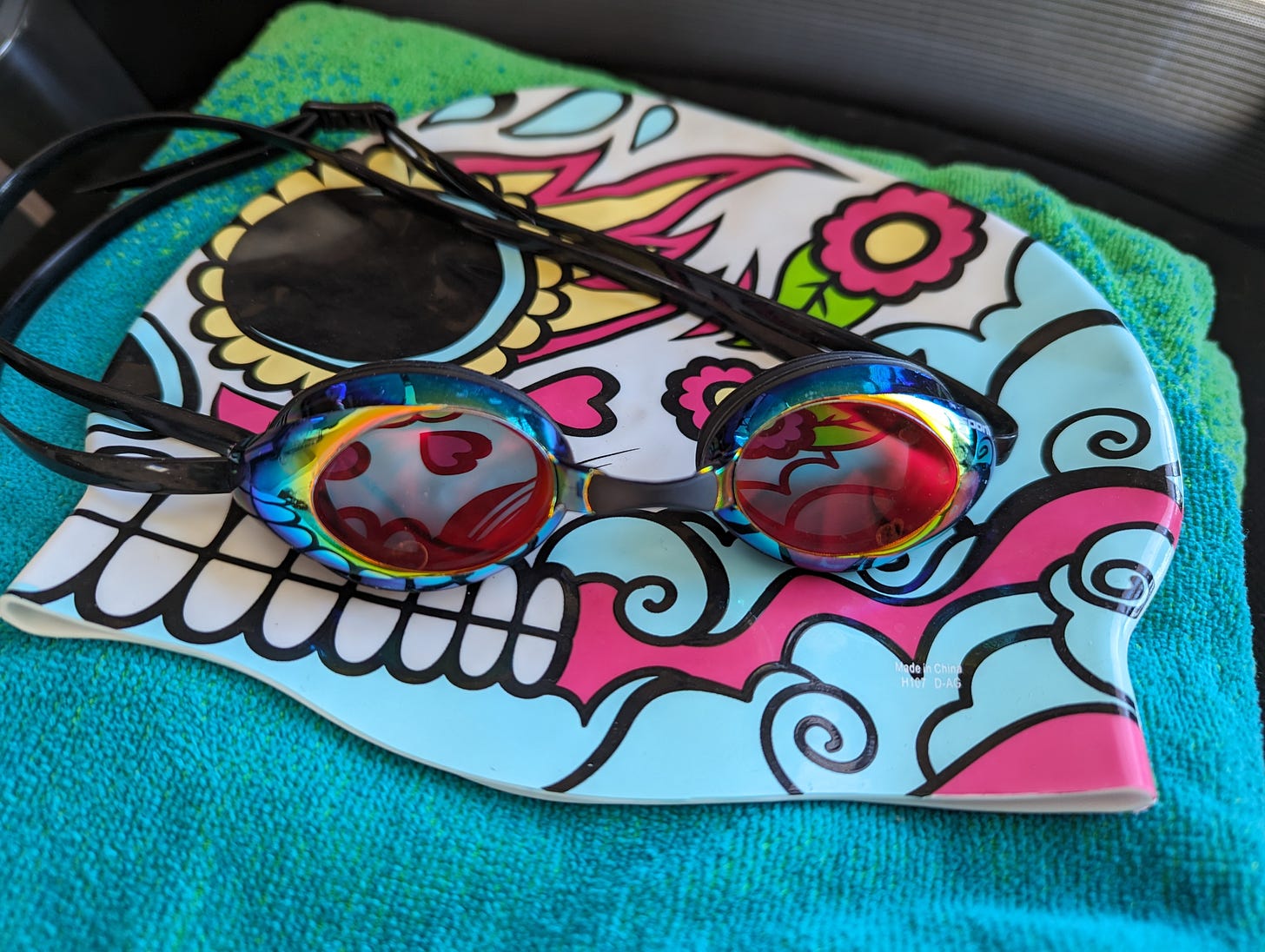 A photo of some of Kate's swim gear: a blue and green beach towel with a sugar skull swim cap on top of it. A pair of rainbow goggles rests on top of the swim cap.