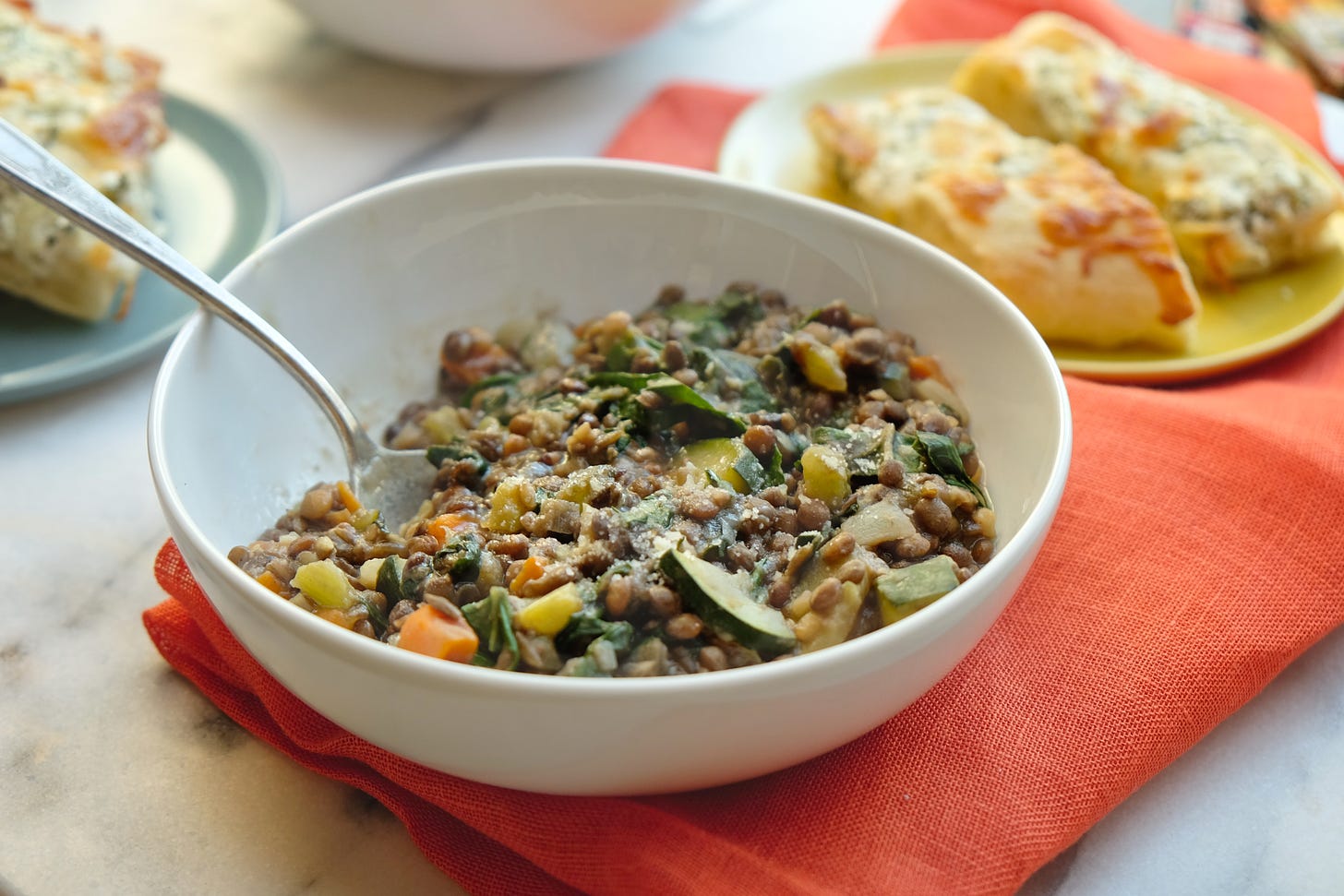 Lentil Stew served in a white bowl set out on top of an orange napkin with a piece of focaccia bread served on a yellow plate to the side