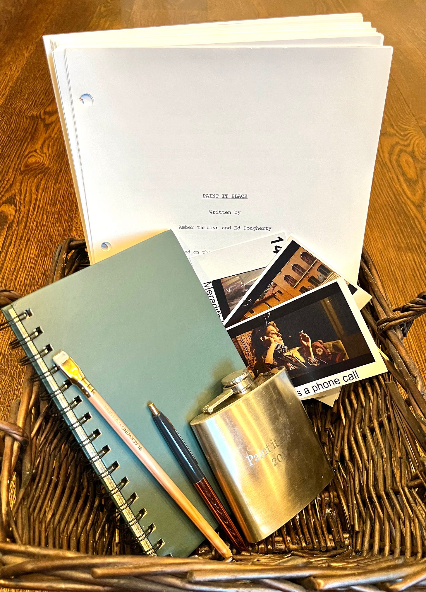 The giveaway bundle. The Paint It Black script, a journal, writing utensils, photos from set, and a flask are arranged in a basket.