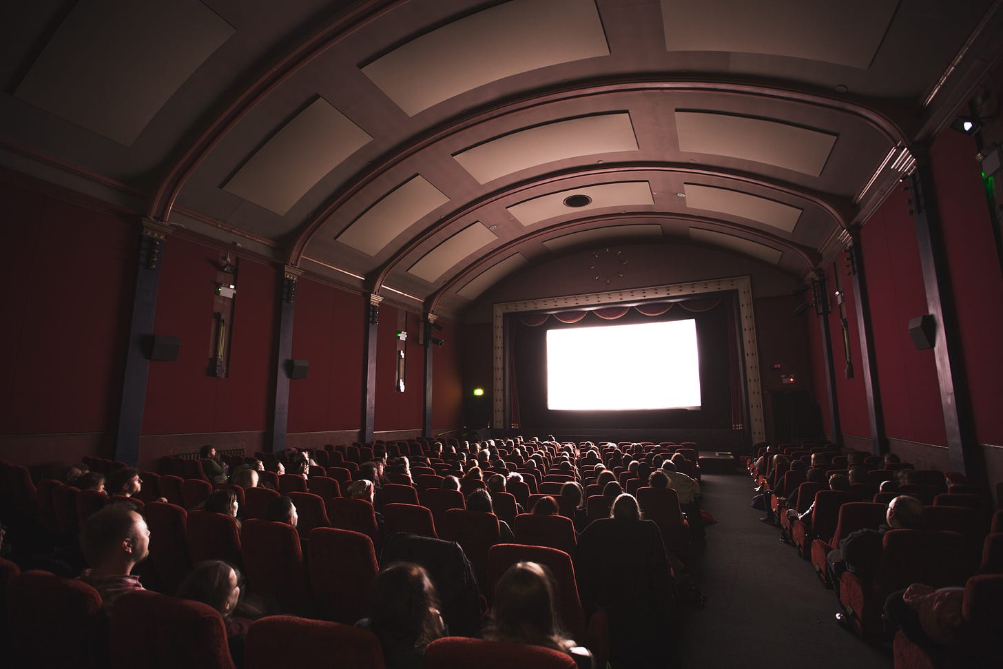 A movie theater mostly full of audience members staring at a blank, white screen. The ceiling of the theater is arched with red velvet walls.