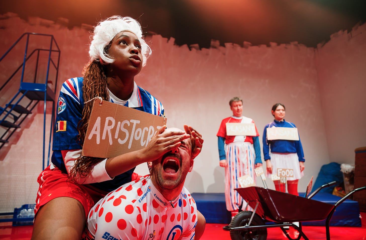 A photograph of The Glorious French Revolution at New Diorama showing an actor wearing a sign saying 'ARISTOCRAT' with their hands over the eyes of another actor who is screaming