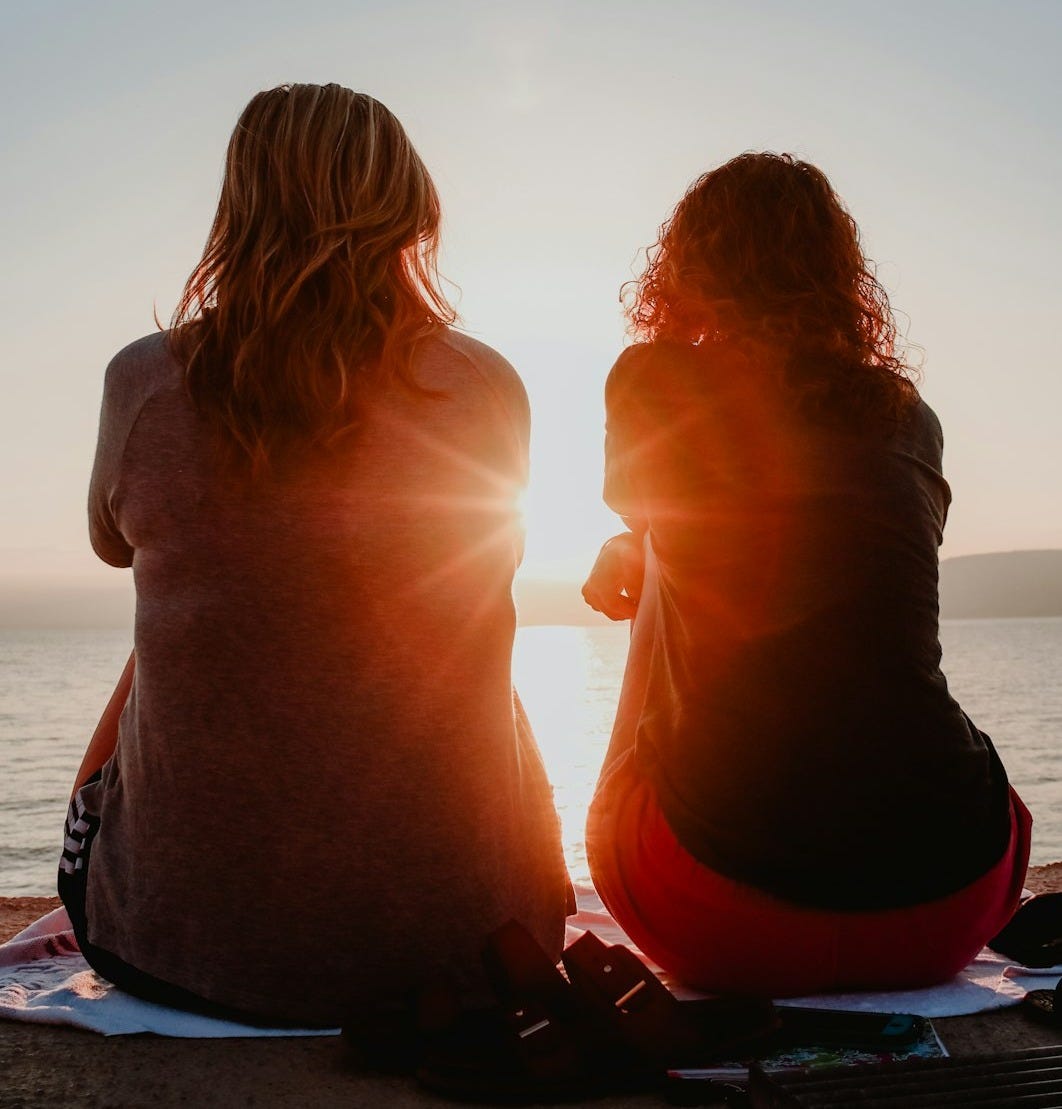 two woman sitting on beach sand while facing sunlight two woman sitting on beach sand while facing sunlight
