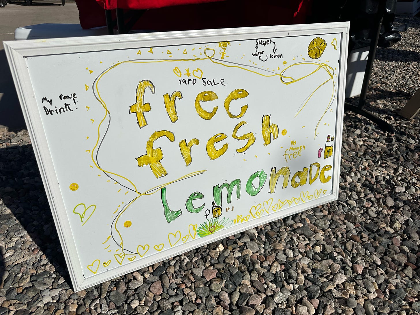 A Libraries and Lemonade sign in front of a stand on a pebbled front yard. It's a white board with hand drawn artwork and the words "Free, fresh, lemonade", "no money free", "my fave drink!" and a diagram showing the relationship between the ingredients (sugar, lemon, and water).