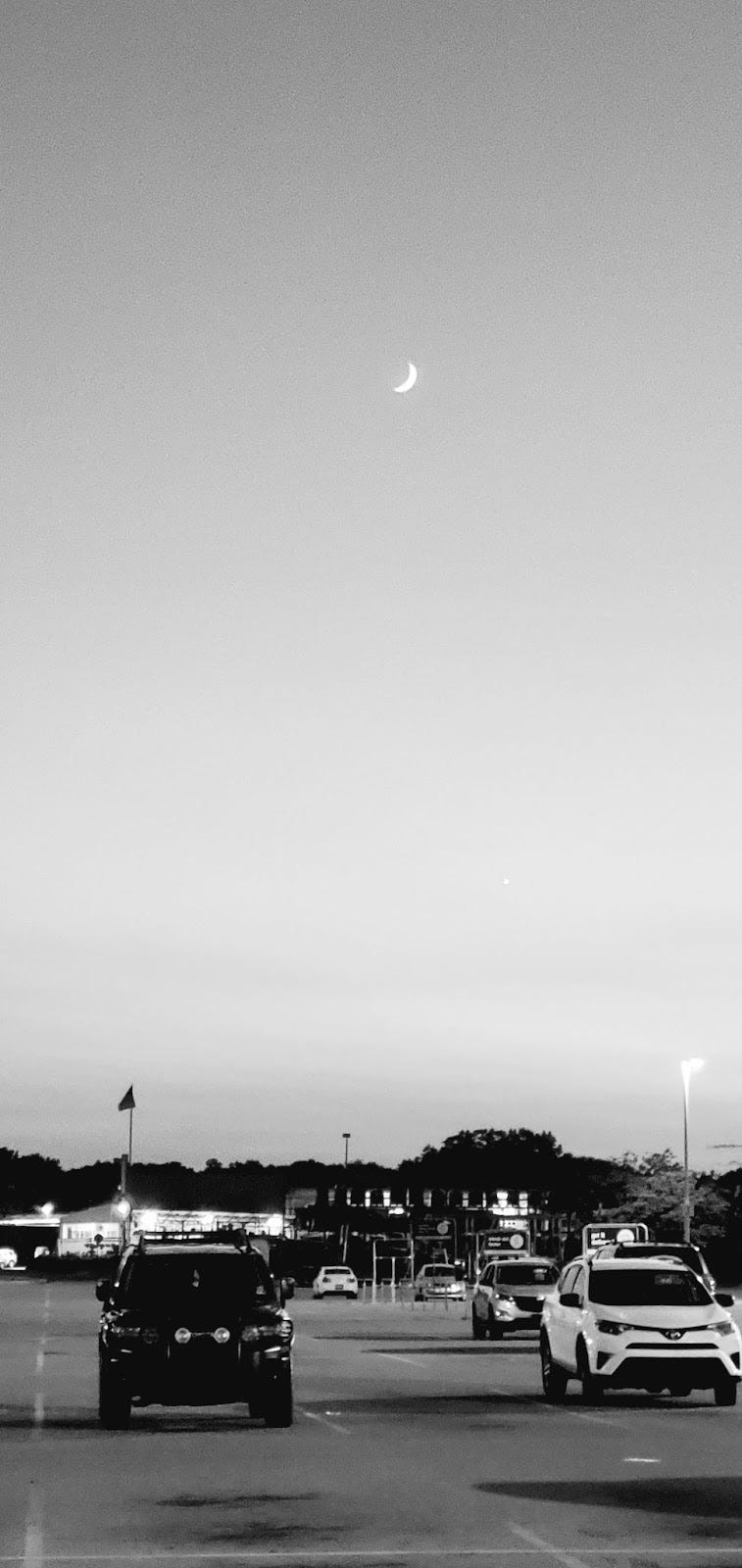 A black-and-white photo of cars on the road, with a large view of the sky and a crescent moon above.