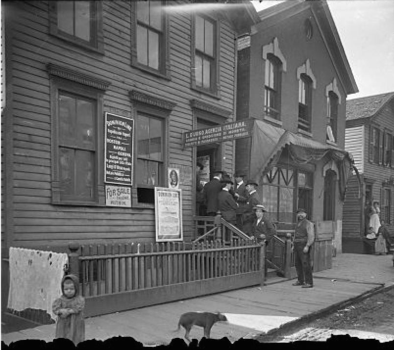 A group of four men on each side of the four stairs leading up to the door. The two men on the one side are facing the two men on the other side and they seem to be talking. One man on the wooden sidewalk and another man on the bottom step are facing the camera. They five men on the steps are wearing suits, the man on the sidewalk is wearing a vest over a long-sleeved shirt. There are signs on the side of the building offering items for sale and services.