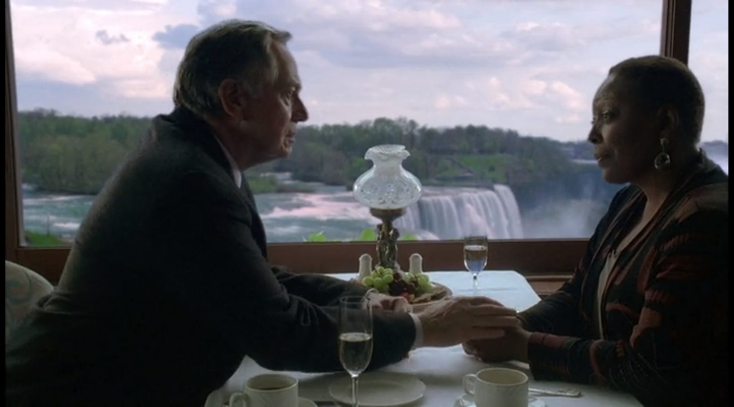 Bernard reaches across a restaurant table to take Rose's hand. Out the window is a real nice view of Niagra Falls.