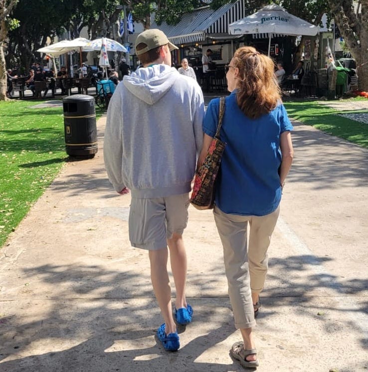 A young man wearing a hoodie, shorts and a ball cap walks with his mother who wears a blue shirt and has long red hair.