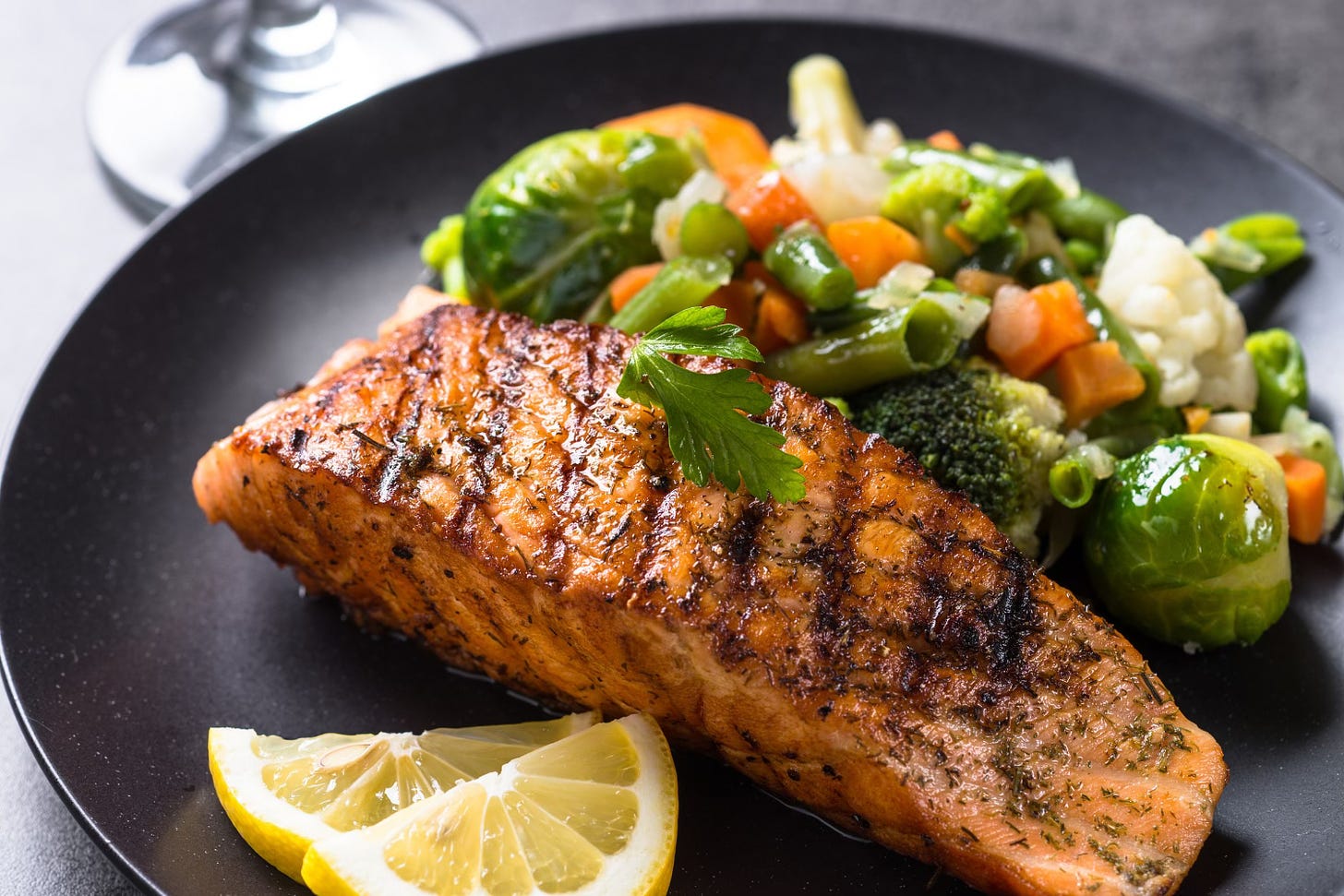 Image of a piece of cooked salmon, some lemon segments and vegetables on a black plate.