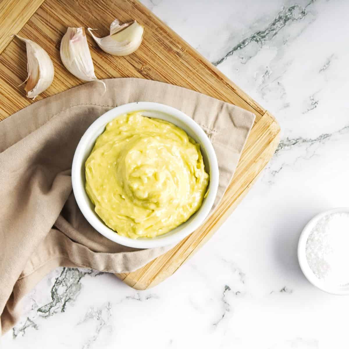 Garlic mayonnaise in small white ramekin on cutting board with tgarlic cloves on marble surface.
