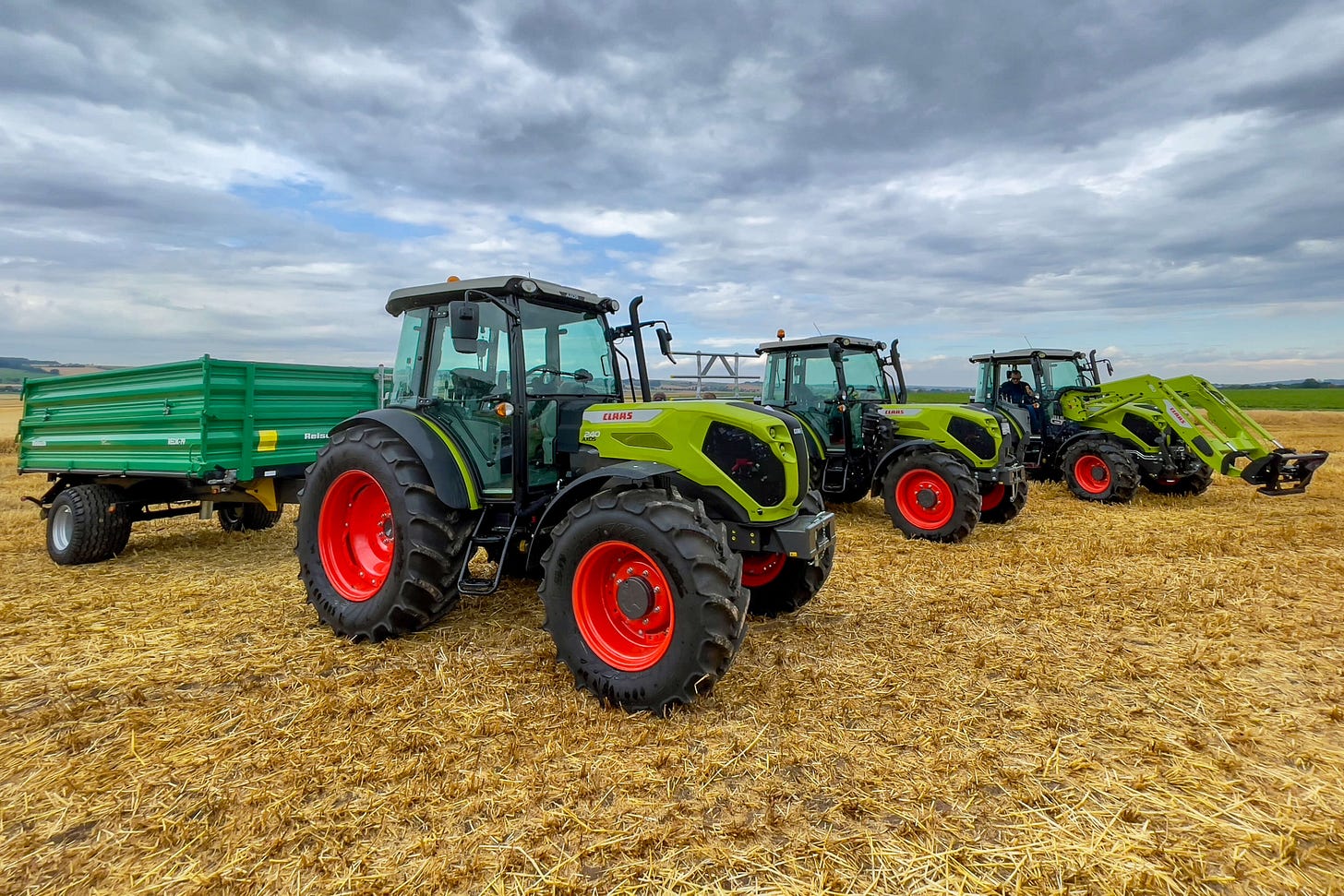 Drei grün-schwarze Claas-Traktoren mit roten Felgen stehen auf einem abgeernteten Feld in einer Reihe. Der vorderste Traktor zieht einen grünen Anhänger, , der hinterste eine Frontladerschwinge. Im Hintergrund ist eine leicht hügelige Landschaft unter bewölktem Himmel zu sehen. Drei grün-schwarze Claas-Traktoren mit roten Felgen stehen auf einem abgeernteten Feld in einer Reihe. Der vorderste Traktor zieht einen grünen Anhänger, , der hinterste eine Frontladerschwinge. Im Hintergrund ist eine leicht hügelige Landschaft unter bewölktem Himmel zu sehen.