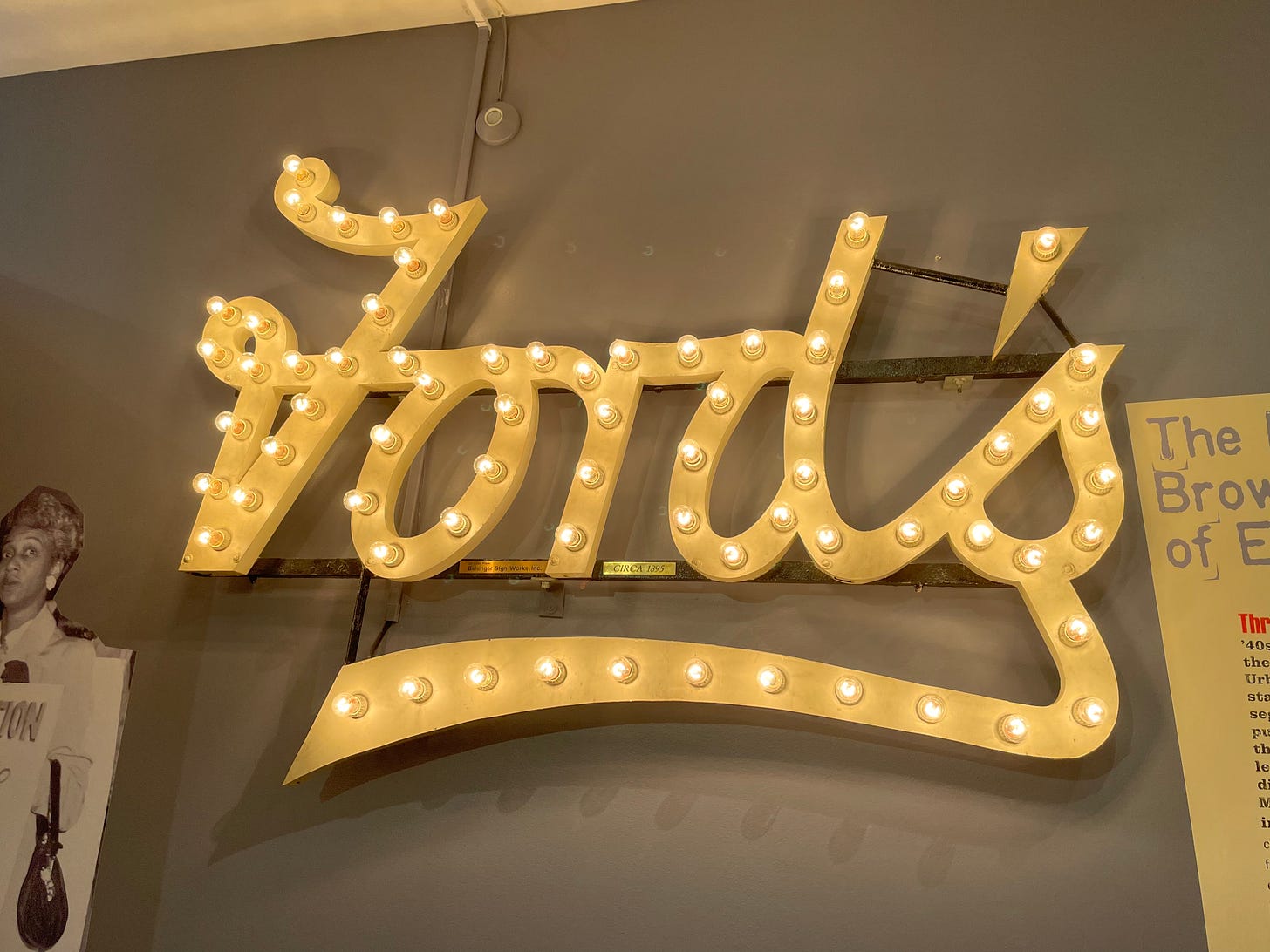 This lighted sign is from the Ford's Grand Opera House in Baltimore City. It spells out "Ford's" in cursive with lightbulbs along the lettering. It is mounted on the wall as part of a museum exhibit.