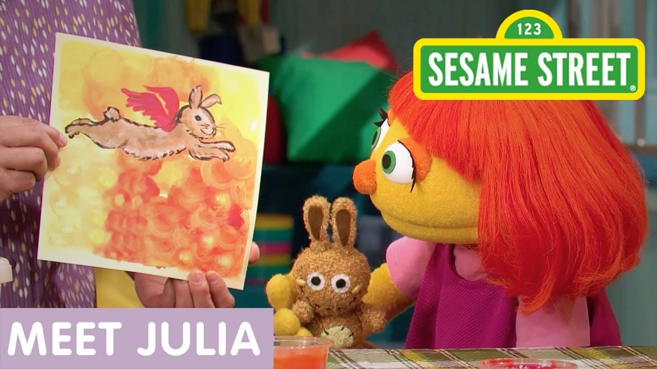 Puppet from Sesame Street named Julia with red hair on the right is holding a stuffed bunny toy while looking at a painting of a bunny