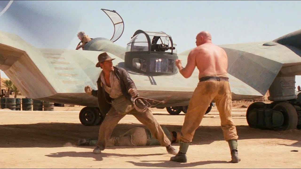 Still from Raiders of the Lost Ark. Indiana Jones winds up to punch the shirtless Nazi bruiser. Behind him, a pilot tries to turn a plane around to catch Jones in the propeller.