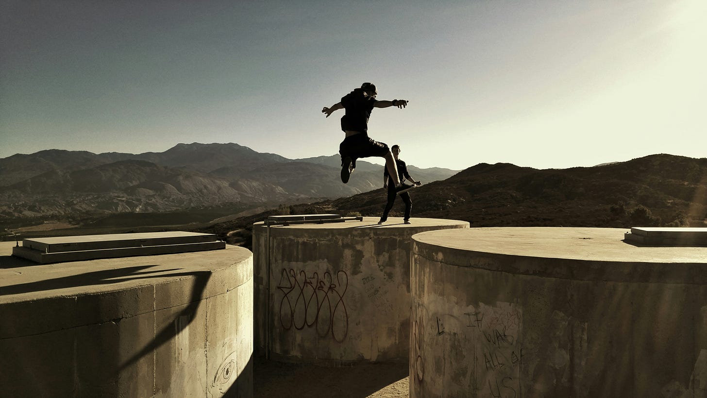 Person leaping across huge concrete containers in sunshine with mountainous backdrop