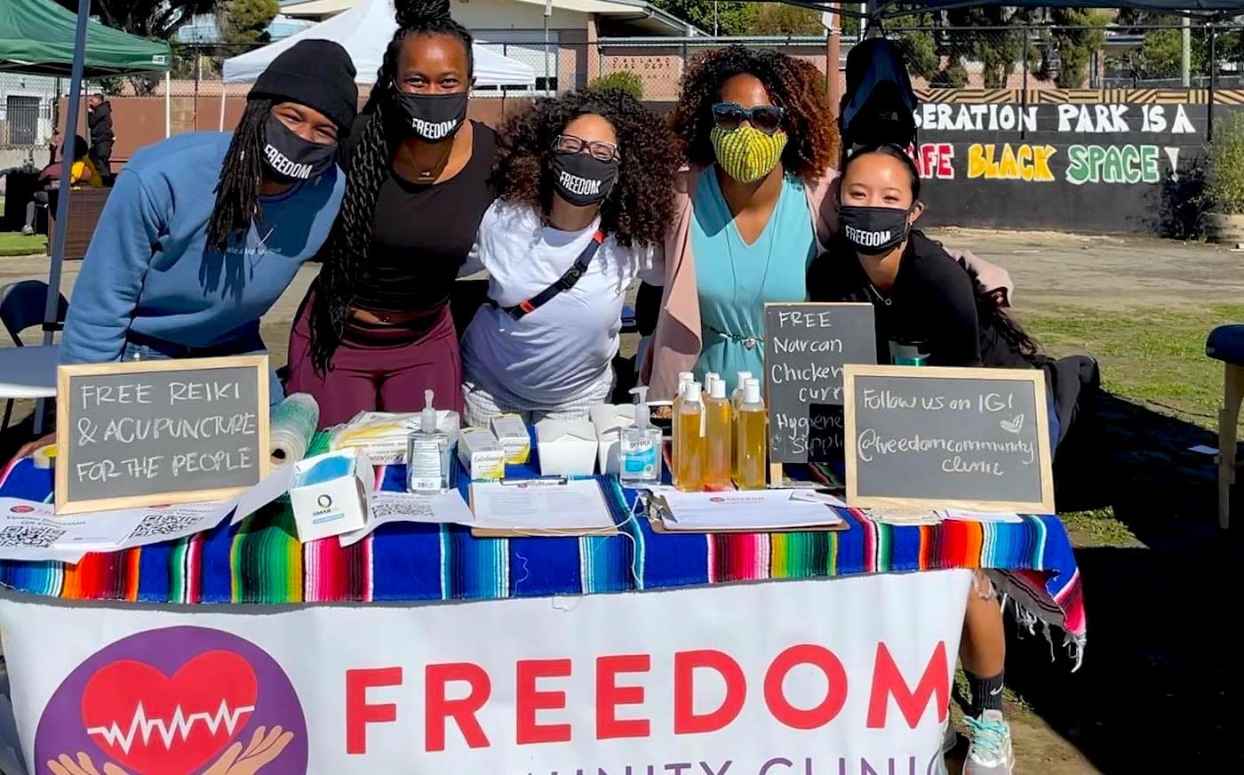 Freedom Community Clinic table at Liberation Park, Bernie and four other team members stand behind it smiling and wearing covid masks that say FREEDOM