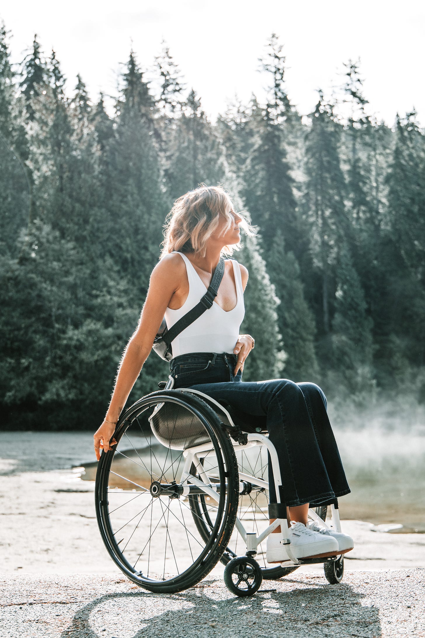 A white woman with long blonde hair sits in a wheelchair, against a backdrop of high top trees with snow around her