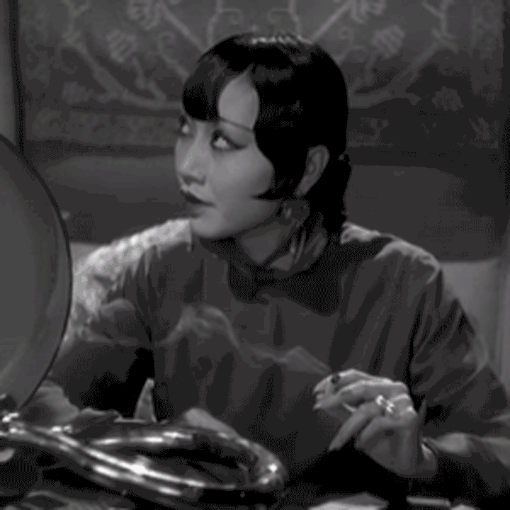 Anna May Wong, as Hui Fei, sitting in a first class train compartment, smokes a cigarette and speaks to someone out of shot to her right. She’s wearing a silk blouse in 1932 film Shanghai Express