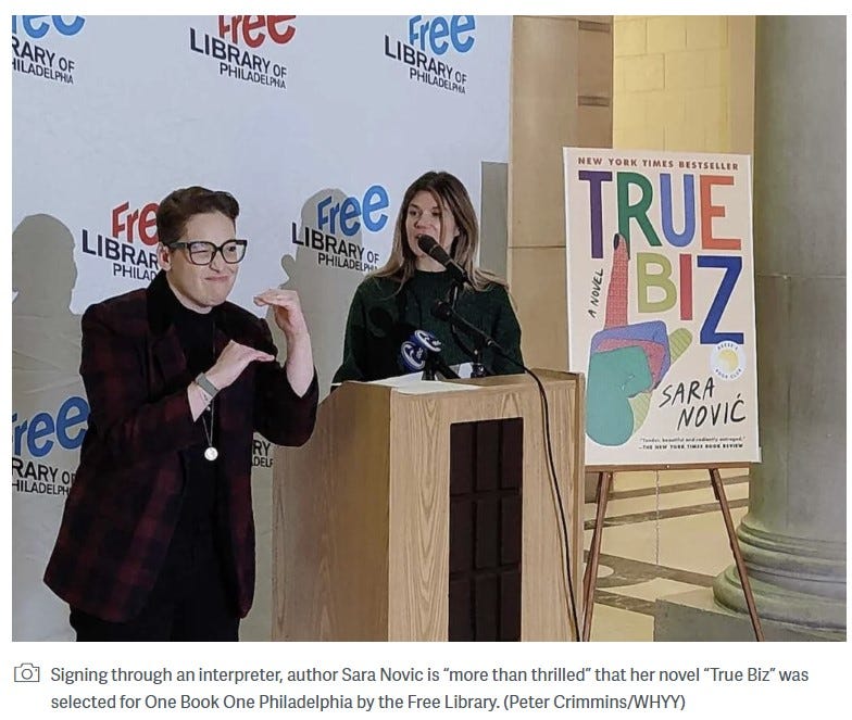 ID: Sara in a red plaid blazer signs “inequity” while the interpreter stands beside her at the podium. A poster of True Biz is on the right side.