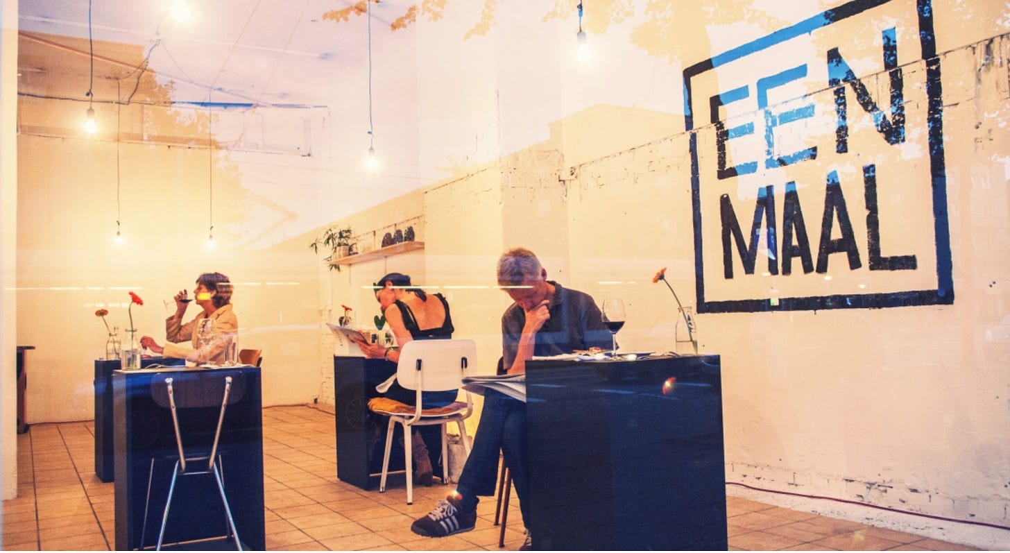 In a glassed-in shop, three diners sit at small black cube tables set for one person each. The words "Een Maal" are painted on a white wall of the space.