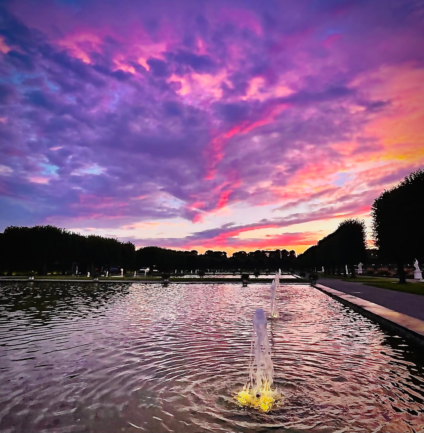A vibrant sunset with deep purples, pinks, and oranges reflecting in the calm water of a fountain pool. The illuminated fountains ripple softly, symbolizing the calm between extremes, with the sky above radiating peaceful energy. Hannover, Herrenhäuser Gärten by Jay Siegmann A vibrant sunset with deep purples, pinks, and oranges reflecting in the calm water of a fountain pool. The illuminated fountains ripple softly, symbolizing the calm between extremes, with the sky above radiating peaceful energy. Hannover, Herrenhäuser Gärten by Jay Siegmann