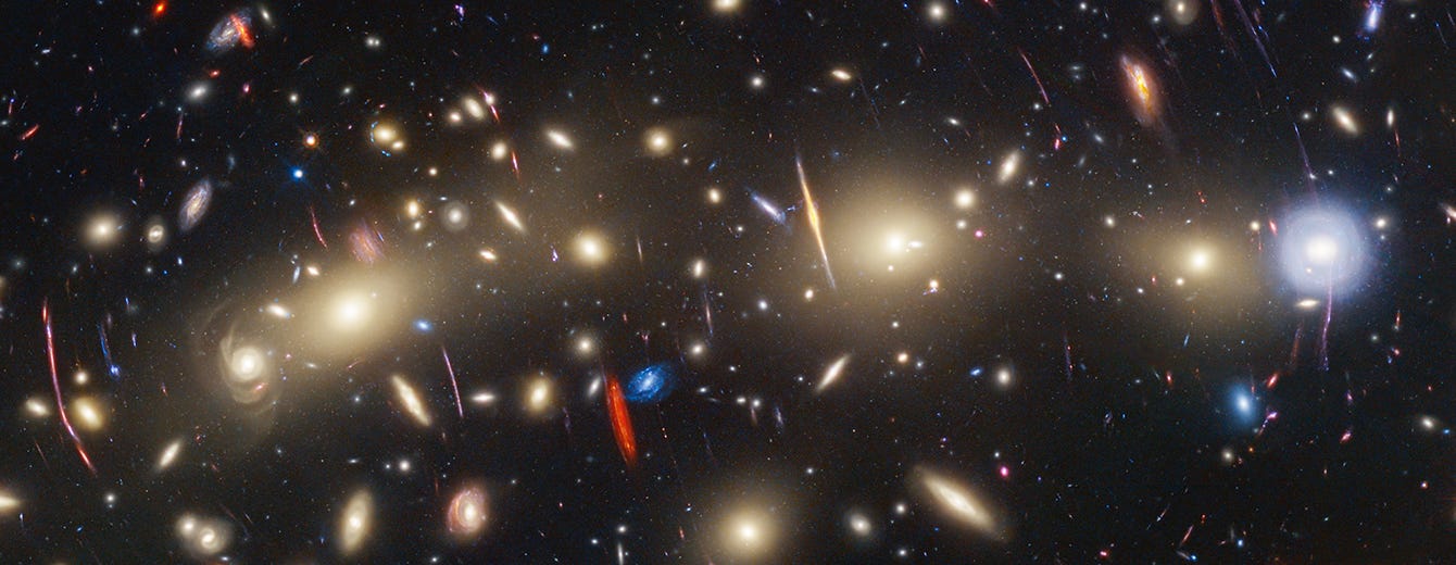 A field of galaxies on the black background of space. Stretching from left to right is a collection of dozens of yellowish spiral and elliptical galaxies that form a foreground galaxy cluster. Among them are distorted linear features, which mostly appear to follow invisible concentric circles curving around the center of the image. The linear features are created when the light of a background galaxy is bent and magnified through gravitational lensing. At left, a particularly prominent example stretches vertically about three times the length of a nearby galaxy. A variety of brightly colored, red and blue galaxies of various shapes are scattered across the image, making it feel densely populated. Near the center are two tiny galaxies compared to the galaxy cluster: a very red edge-on spiral and a very blue face-on spiral, which provide a striking color contrast.