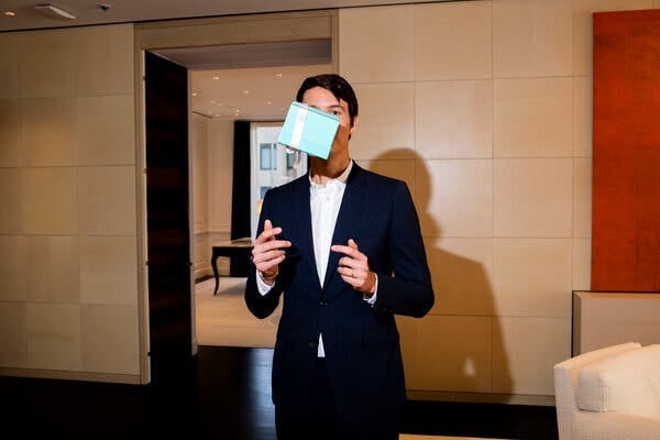 A man in a blue suit, with a white shirt and no necktie, is pictured standing in a room at Tiffany with a light blew jewelry box at the level of his head. He has apparently just tossed the box into the air.