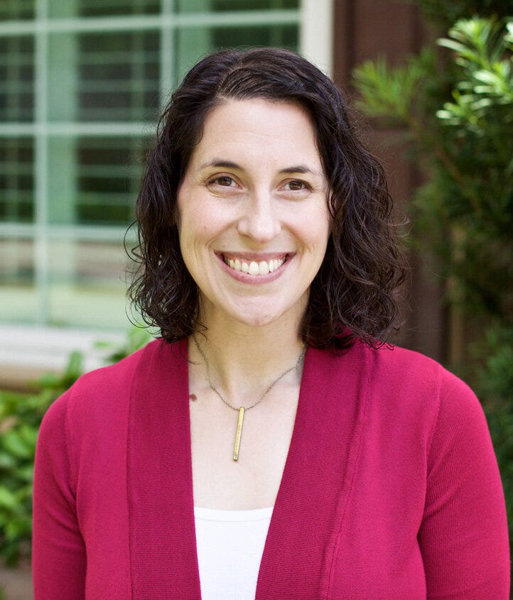 rebecca is a white-skinned woman with shoulder length brown hair. she wears a white blouse and magenta jacket.she smiles at the camera